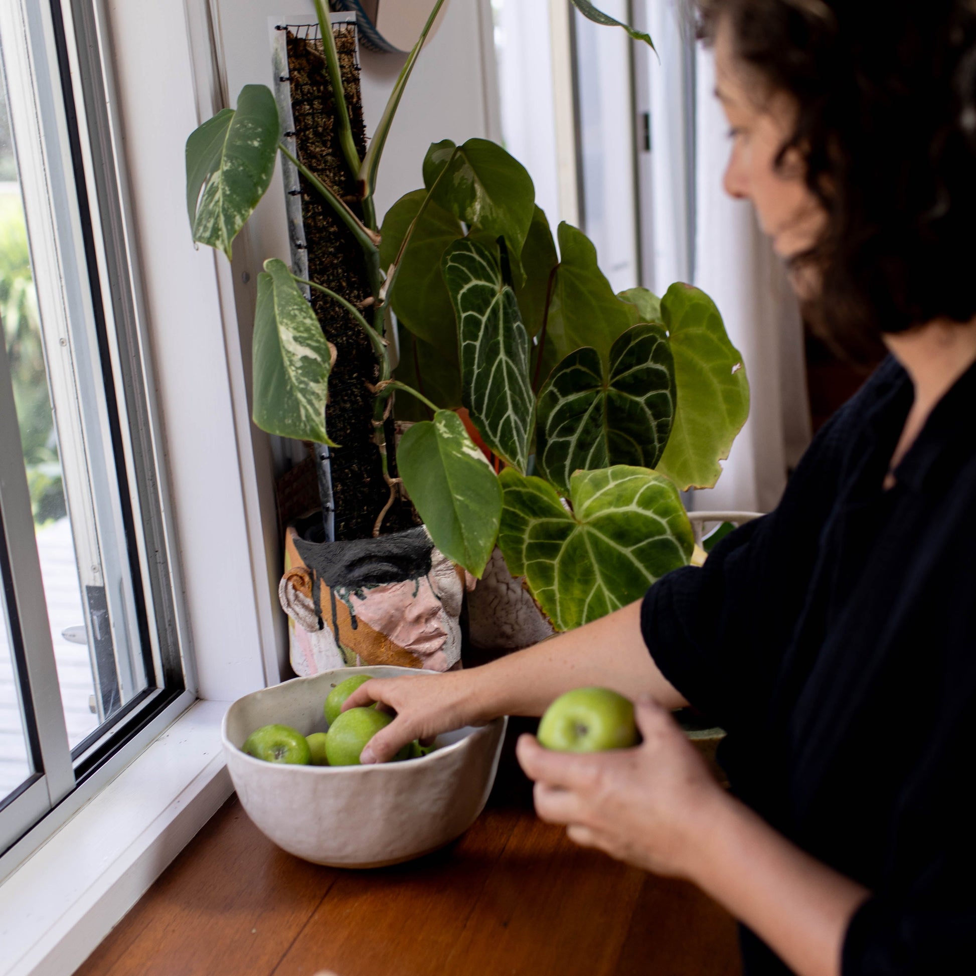 Person selecting apples from a bowl on a wooden table with a plant in the background