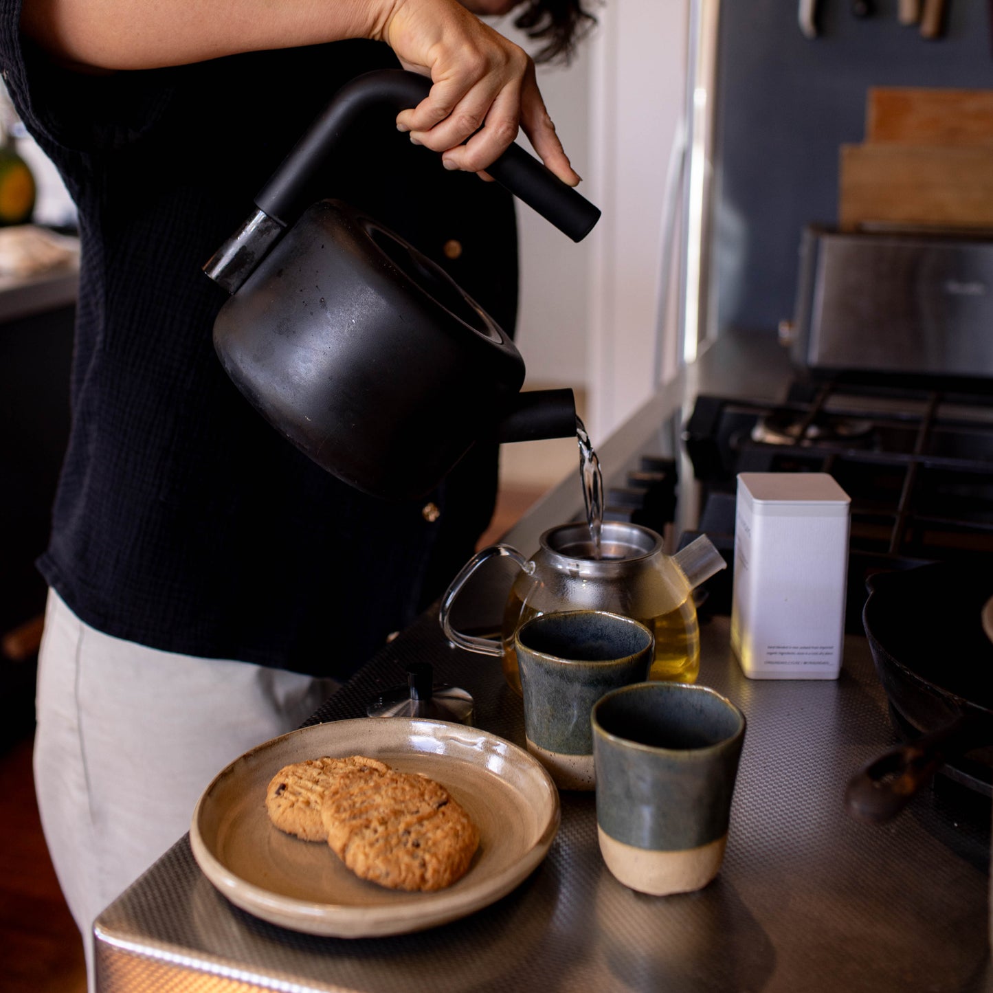 Person pouring from a black teapot into two cups on a kitchen counter.