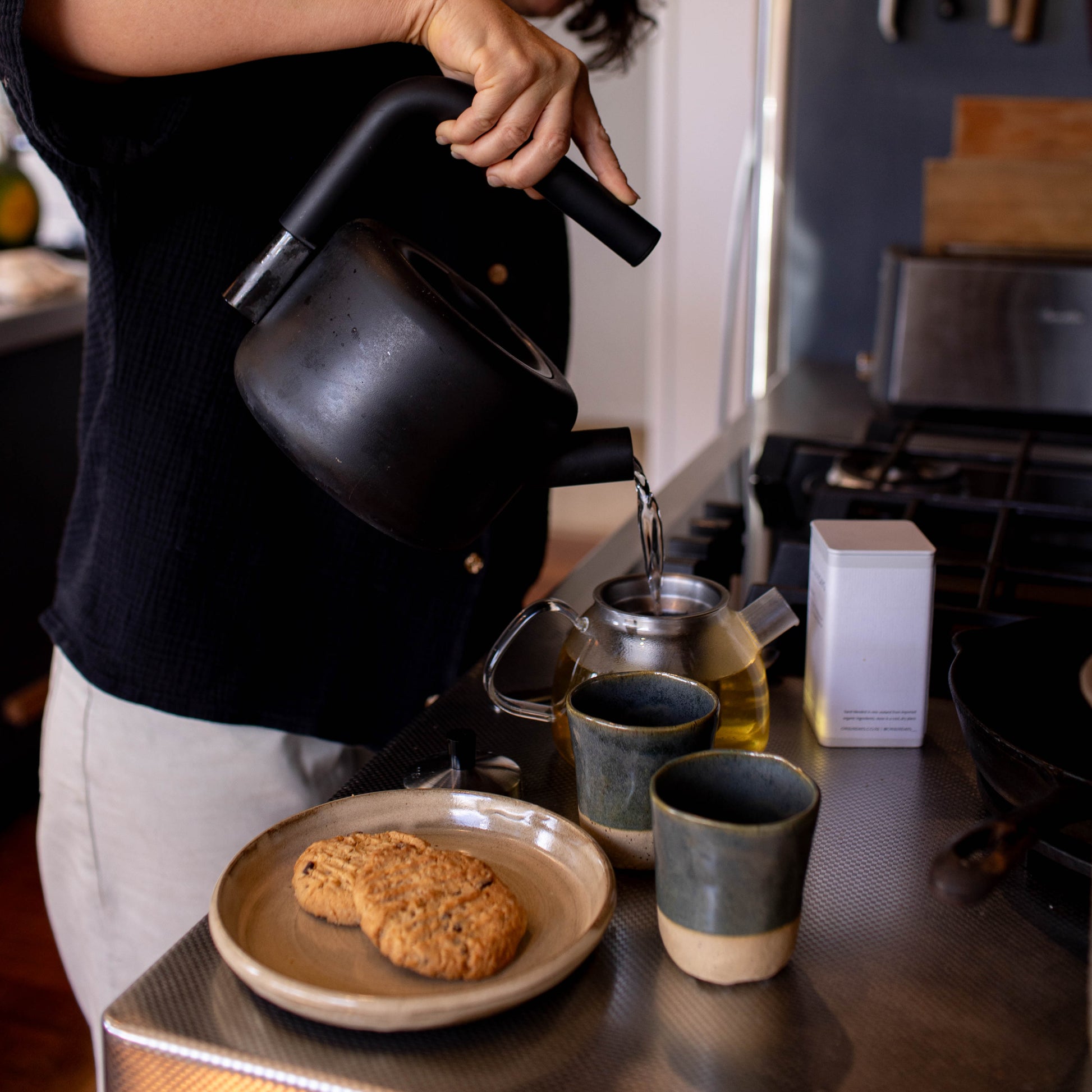 Person pouring from a black teapot into two cups on a kitchen counter.