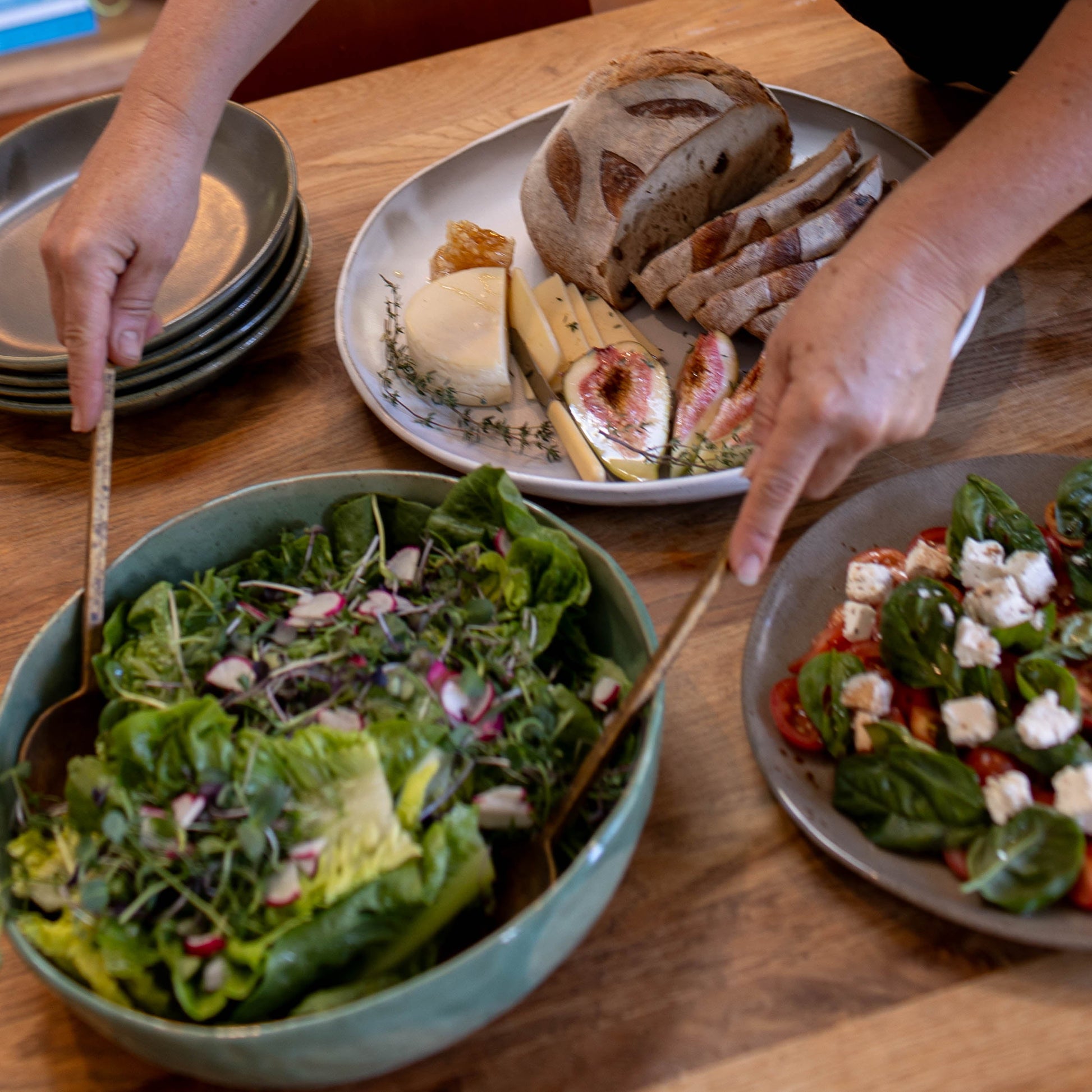 Person preparing a meal with salad, bread, and cheese on a wooden table.
